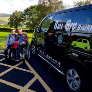 two people, one crowching beside person in wheelchair, beside a black car